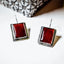 Pair of square silver drop earrings with deep red carnelian stones in a double-edged silver frame, displayed on a white surface with a silver pine cone in the background.