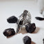Large sculptural silver ring with a crumpled free-form polished surface showing dramatic light and shadow between the folded planes, displayed among black rocks on a white surface.