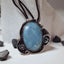 Large oval chaolite pendant in a sky blue tone set in an ornate oxidised silver frame with scroll details and silver ball accents, on a dark brown cord leaning against a stone surface.