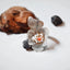 Silver flower ring with four cupped textured petals and a cluster of white seed pearls and a pink coral bead at the centre, leaning against a pine cone and dark pebble on a white surface.