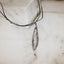 Long slender oxidised silver olive leaf pendant with a deeply textured surface, hanging on a black cord against a white marble surface.