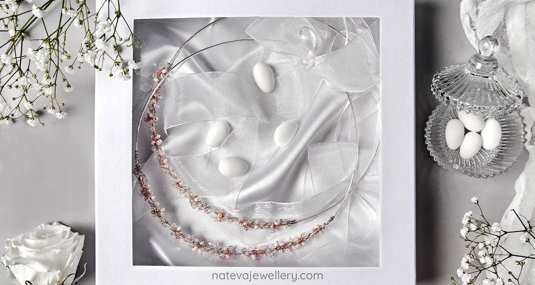 Elegant presentation of handmade Greek wedding crowns (stefana) in a white gift box with satin fabric, surrounded by white roses, sugared almonds and bridal accents.