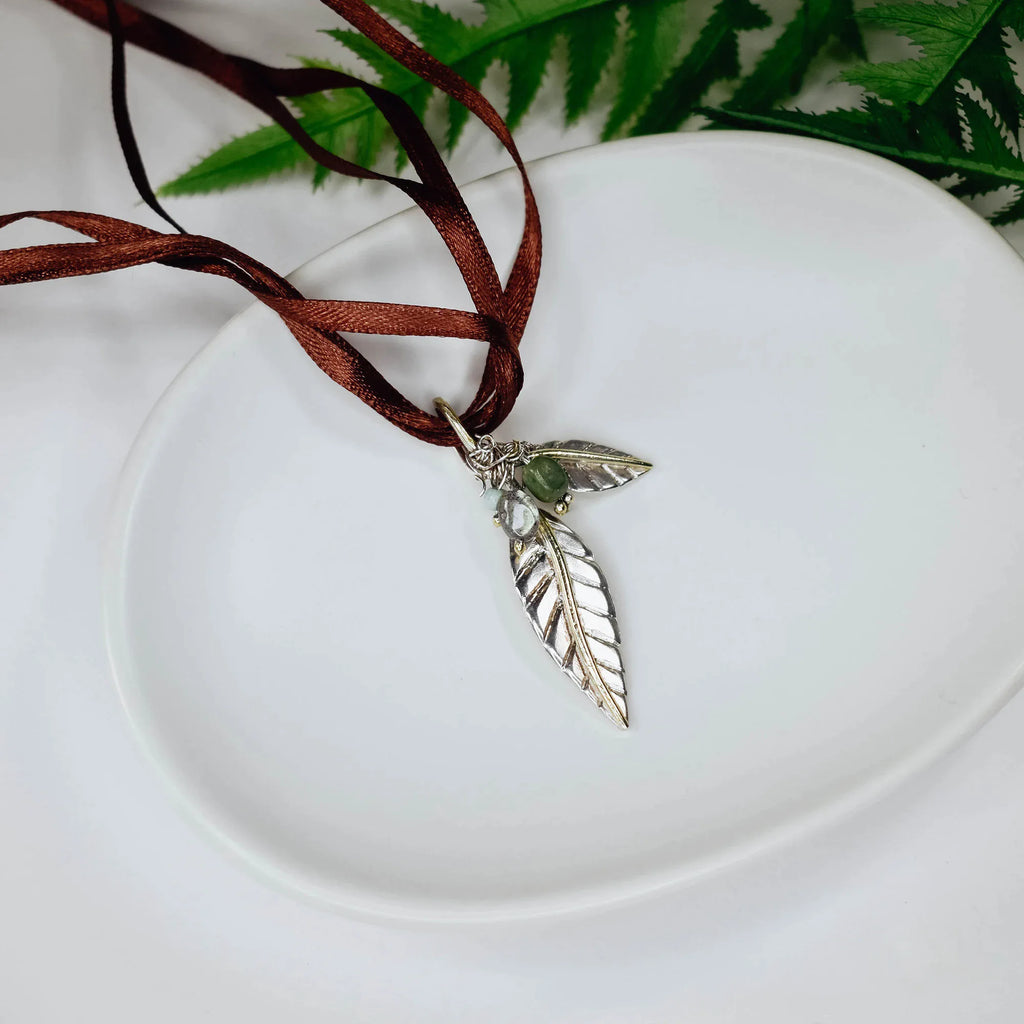 Silver leaf pendant featuring two leaves and a wire-wrapped cluster of moonstone, turquoise and green gemstone, on a brown satin ribbon cord displayed on a white plate.