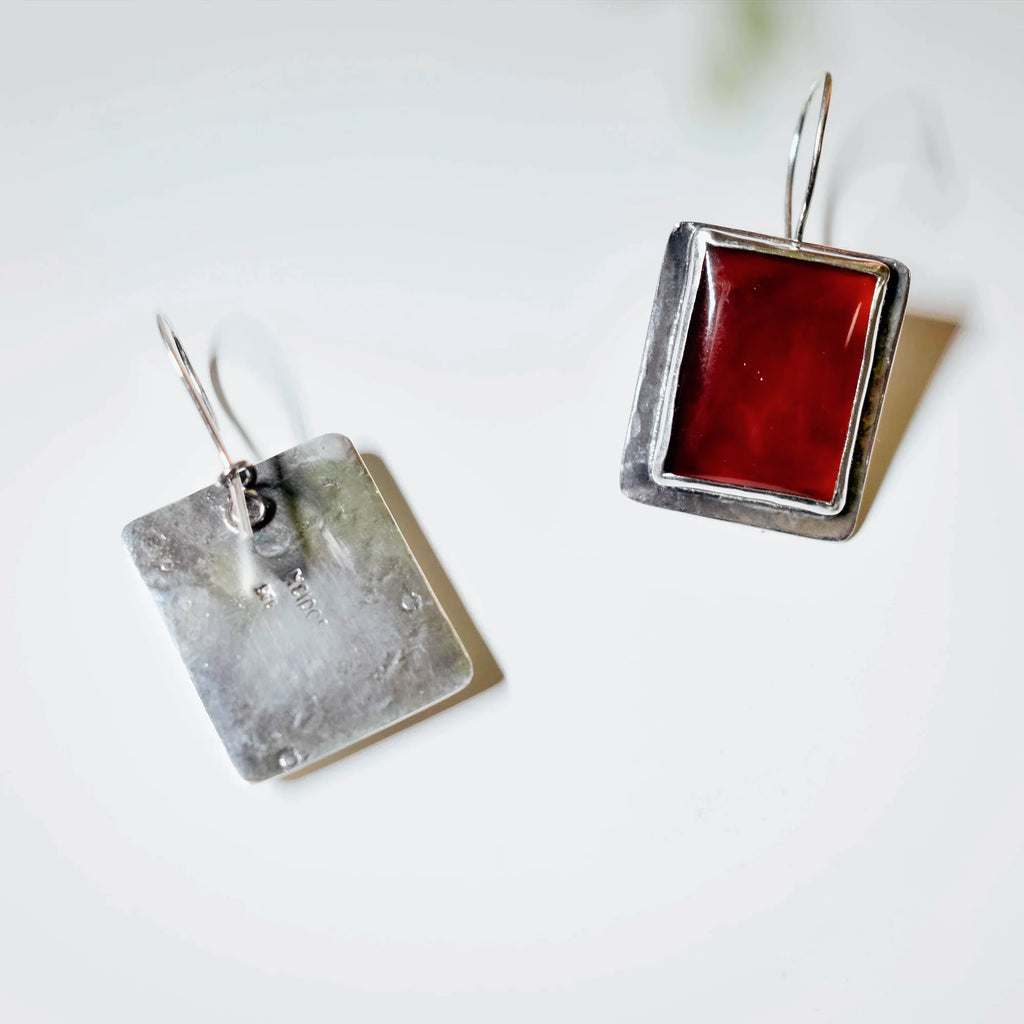 Back and front view of square silver drop earrings showing the plain silver back of one earring and the deep red carnelian front setting of the other, on a white surface.