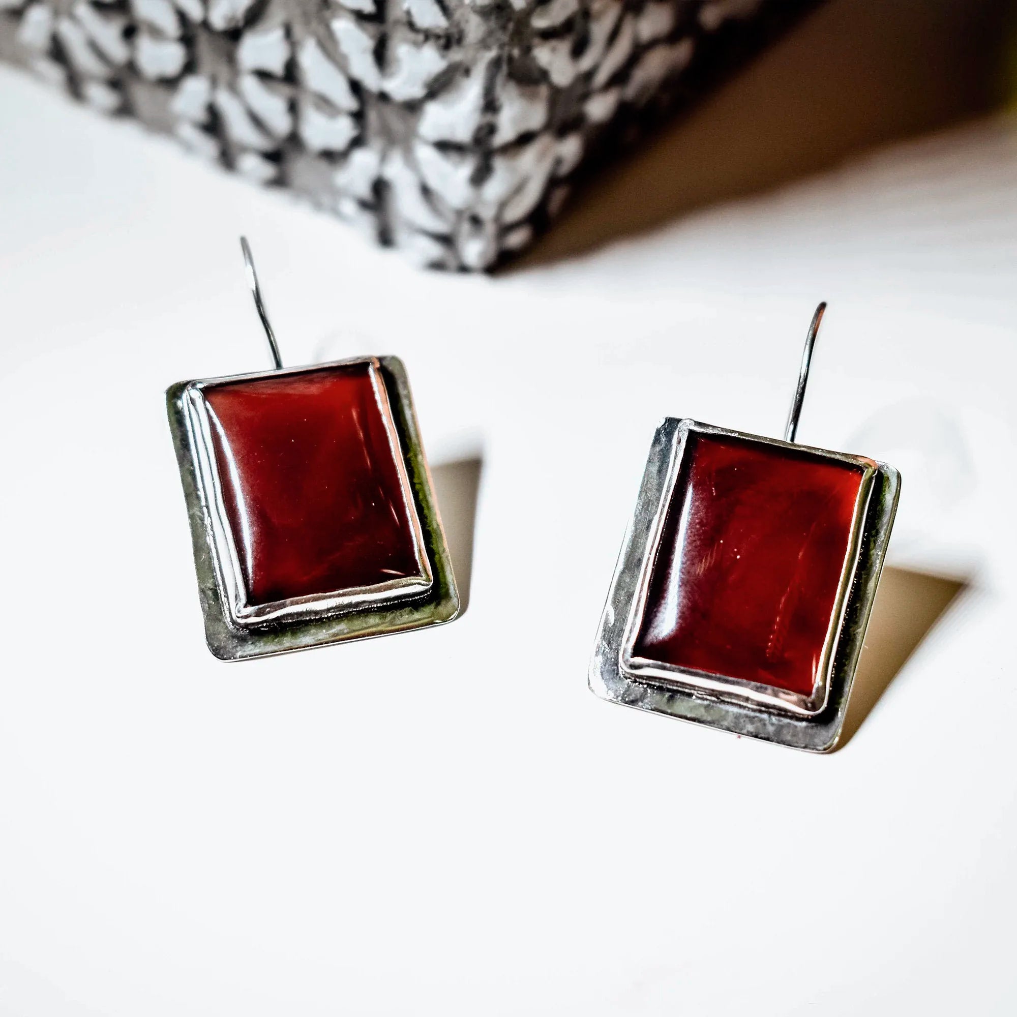Pair of square silver drop earrings with deep red carnelian stones in a double-edged silver frame, displayed on a white surface with a silver pine cone in the background.