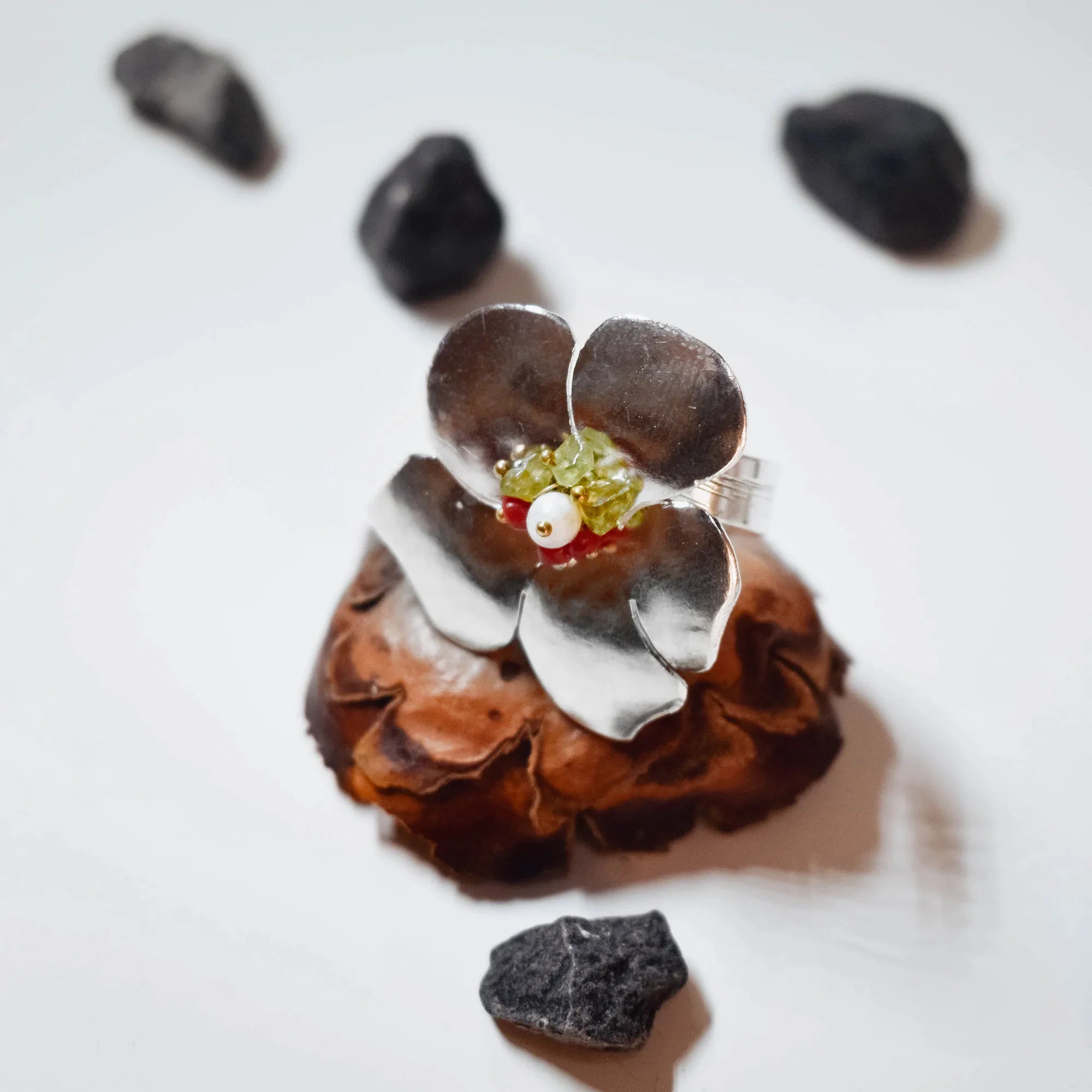 Large silver flower ring with four brushed petals and a centre cluster of green crystal chips, red coral, white freshwater pearl and gold beads, displayed on a pine cone with black stones around it. Main image