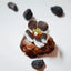 Large silver flower ring with four brushed petals and a centre cluster of green crystal chips, red coral, white freshwater pearl and gold beads, displayed on a pine cone with black stones around it.