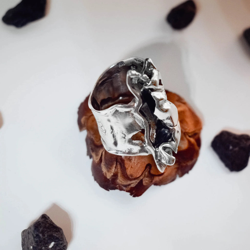 Overhead view of a large sculptural silver ring showing the wild free-form crumpled silver top and the open adjustable band on a pine cone with black stones.a