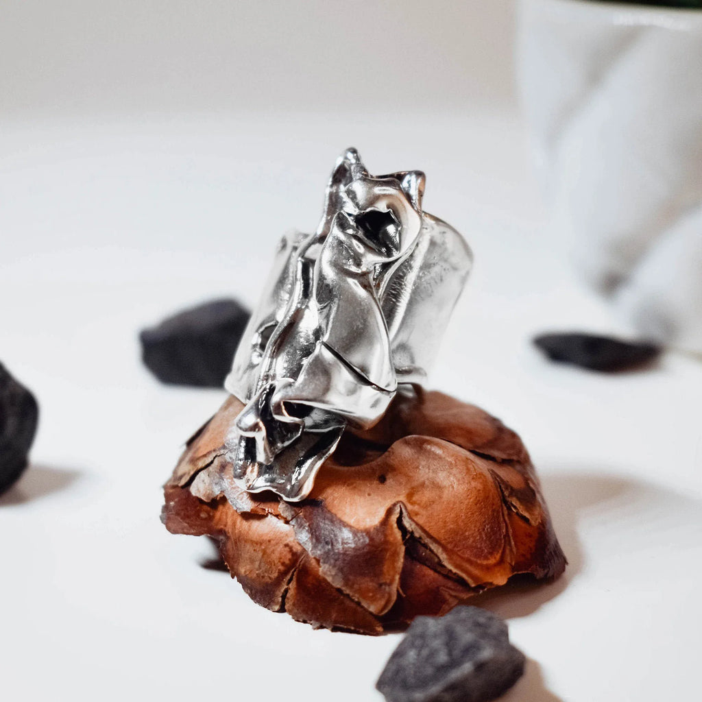  Large sculptural silver ring with a dramatic crumpled free-form polished surface displayed on a pine cone with black stones around it, side view.