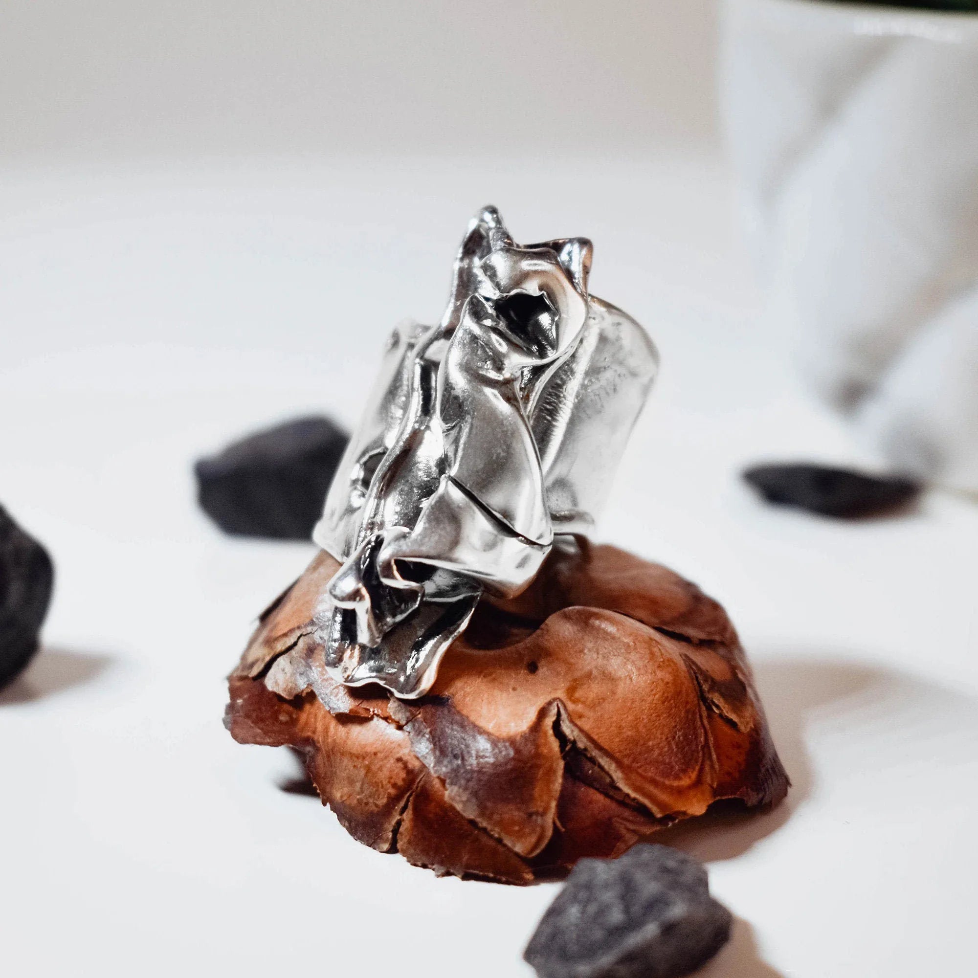 Large sculptural silver ring with a dramatic crumpled free-form polished surface displayed on a pine cone with black stones around it, side view. Main image