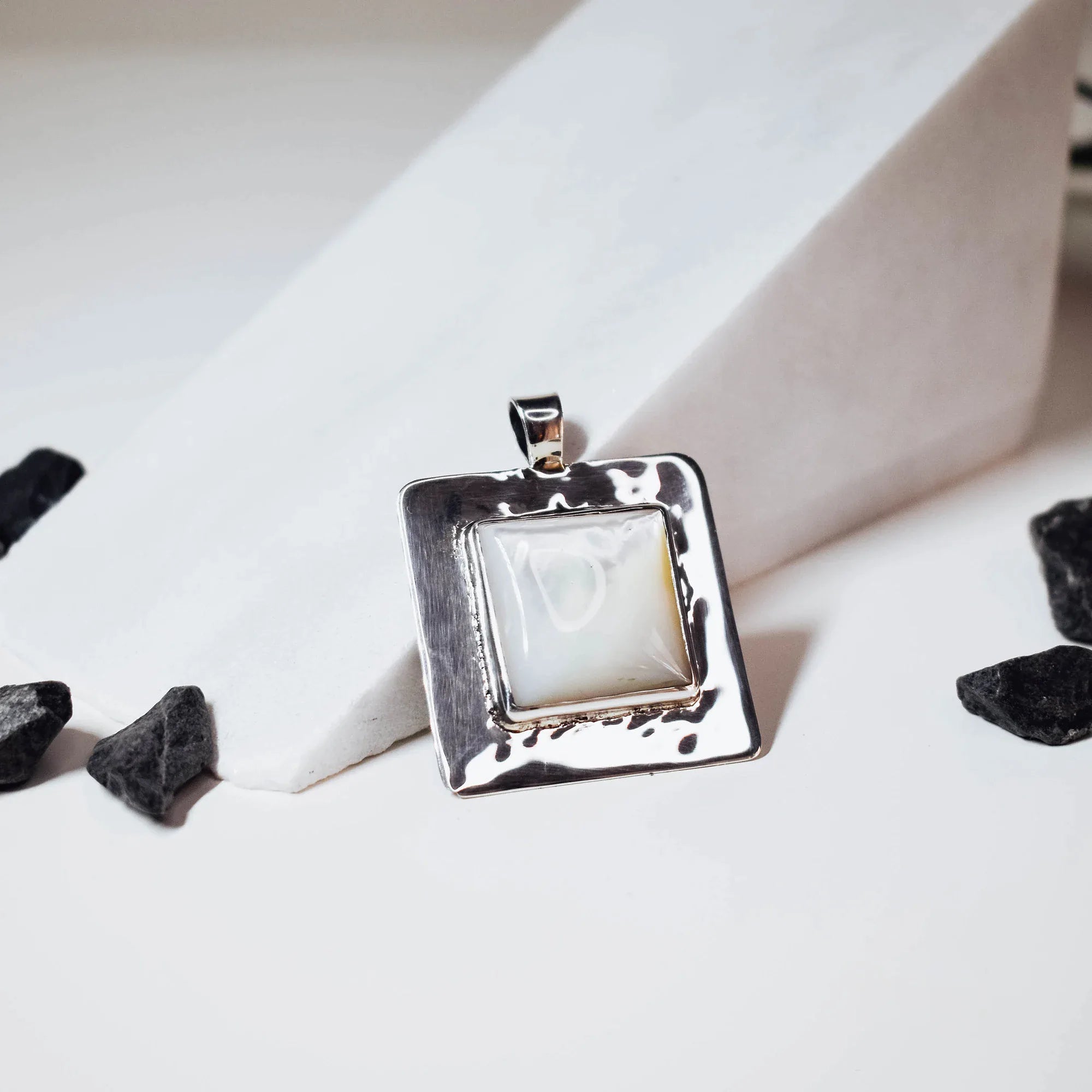 Square hammered silver pendant with softly rounded corners and a smooth fildisi cabochon centre stone, displayed against a white surface with black stones around it. Main image