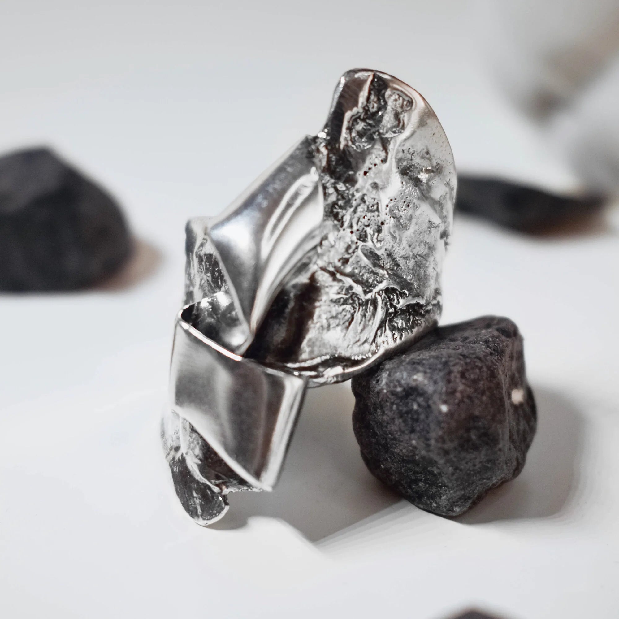 Side view of a sculptural silver ring showing the folded and crumpled high-polish silver surface with natural shadow contrast between the planes, among black rocks.