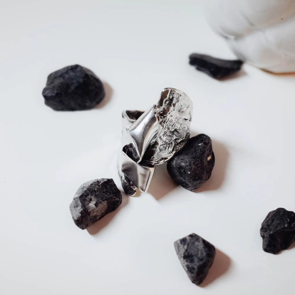 Front view of a sculptural silver ring showing the full crumpled polished silver surface surrounded by black rocks on a white surface.