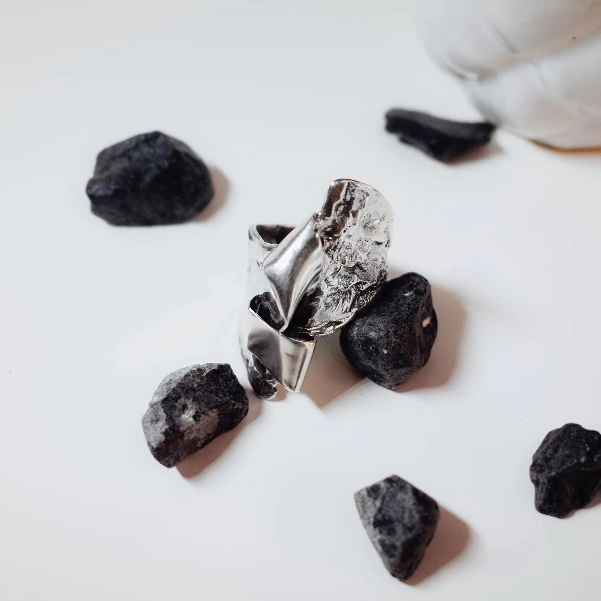 Front view of a sculptural silver ring showing the full crumpled polished silver surface surrounded by black rocks on a white surface.