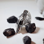 Large sculptural silver ring with a crumpled free-form polished surface showing dramatic light and shadow between the folded planes, displayed among black rocks on a white surface.