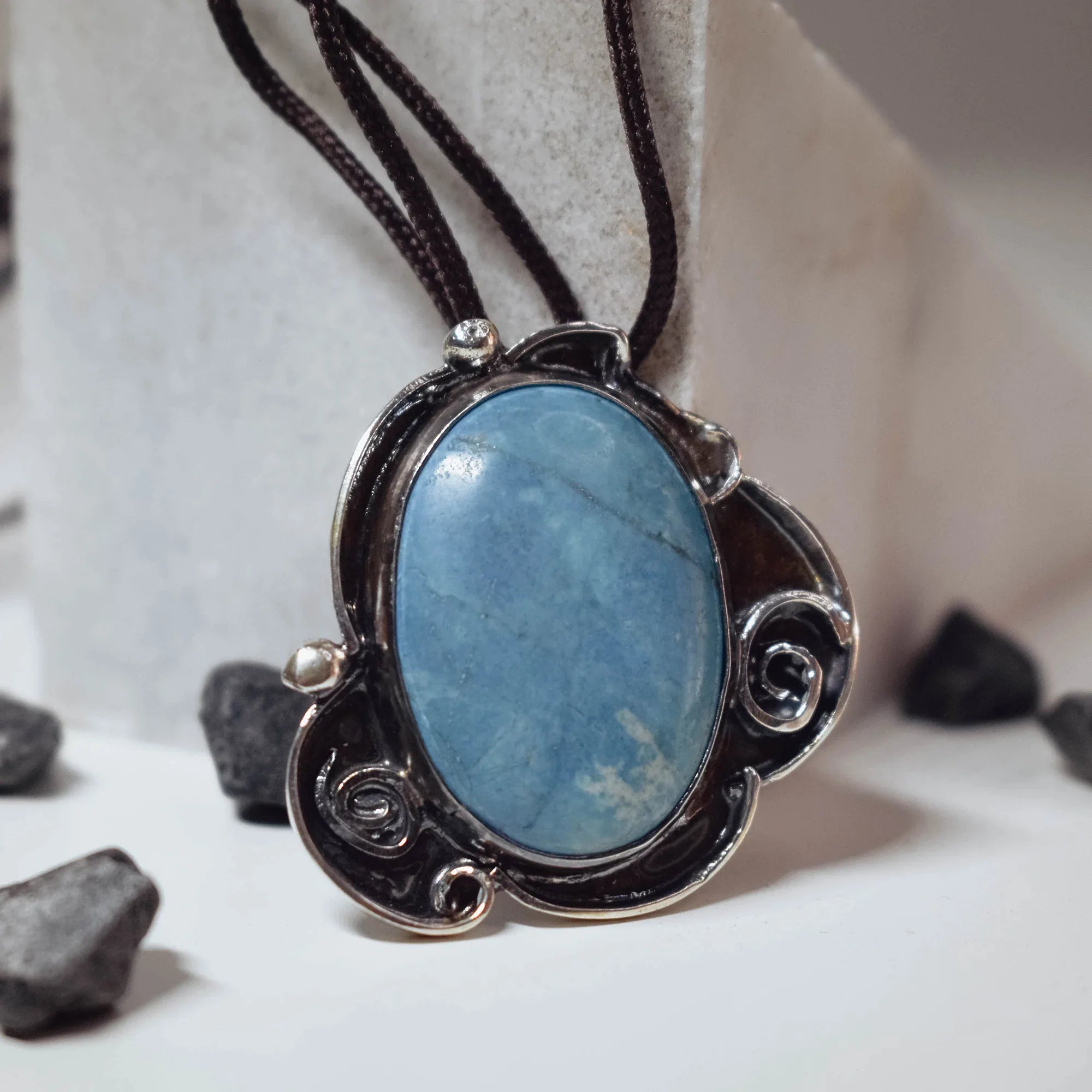 Large oval chaolite pendant in a sky blue tone set in an ornate oxidised silver frame with scroll details and silver ball accents, on a dark brown cord leaning against a stone surface.
