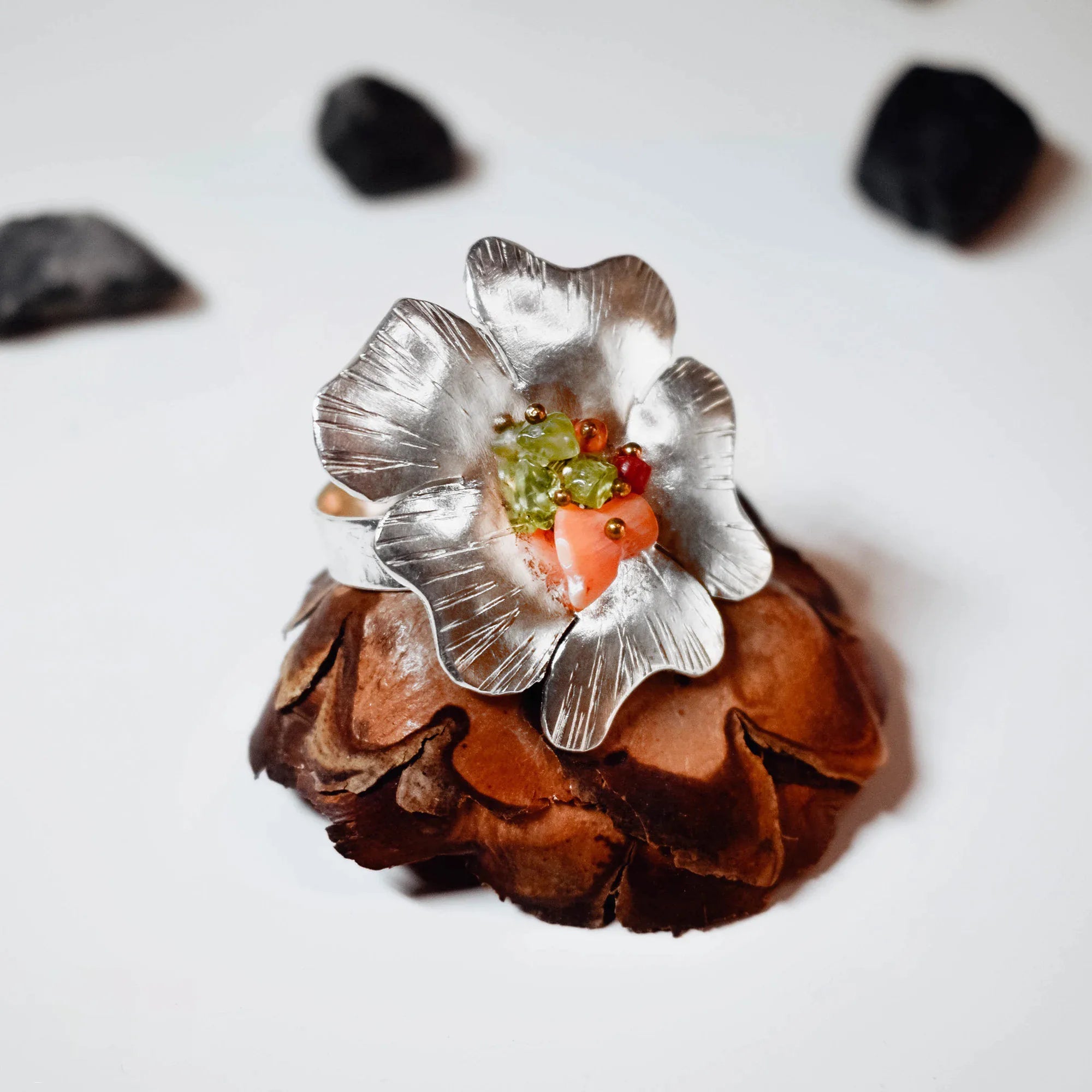 Top view of a large silver flower ring showing the full brushed petal detail and the warm-toned gemstone centre cluster of green tourmaline and coral on a pine cone display.