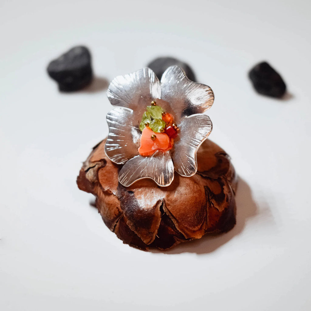 Large silver flower ring with brushed petals and a centre cluster of green tourmaline chips, coral chips and gold beads, displayed on a pine cone with black stones around it.