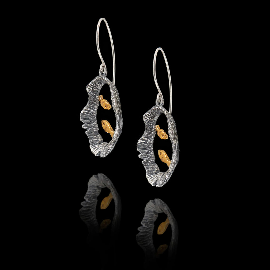 Angled side view of a pair of silver earrings with gold fish design on a black background