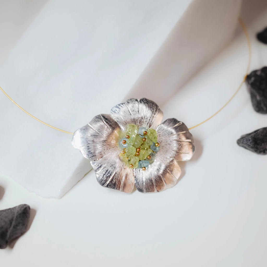 Close-up of a large sculpted silver flower pendant showing the detailed brushed petal texture and raw green tourmaline chip centre with tiny gold beads on a gold wire choker.