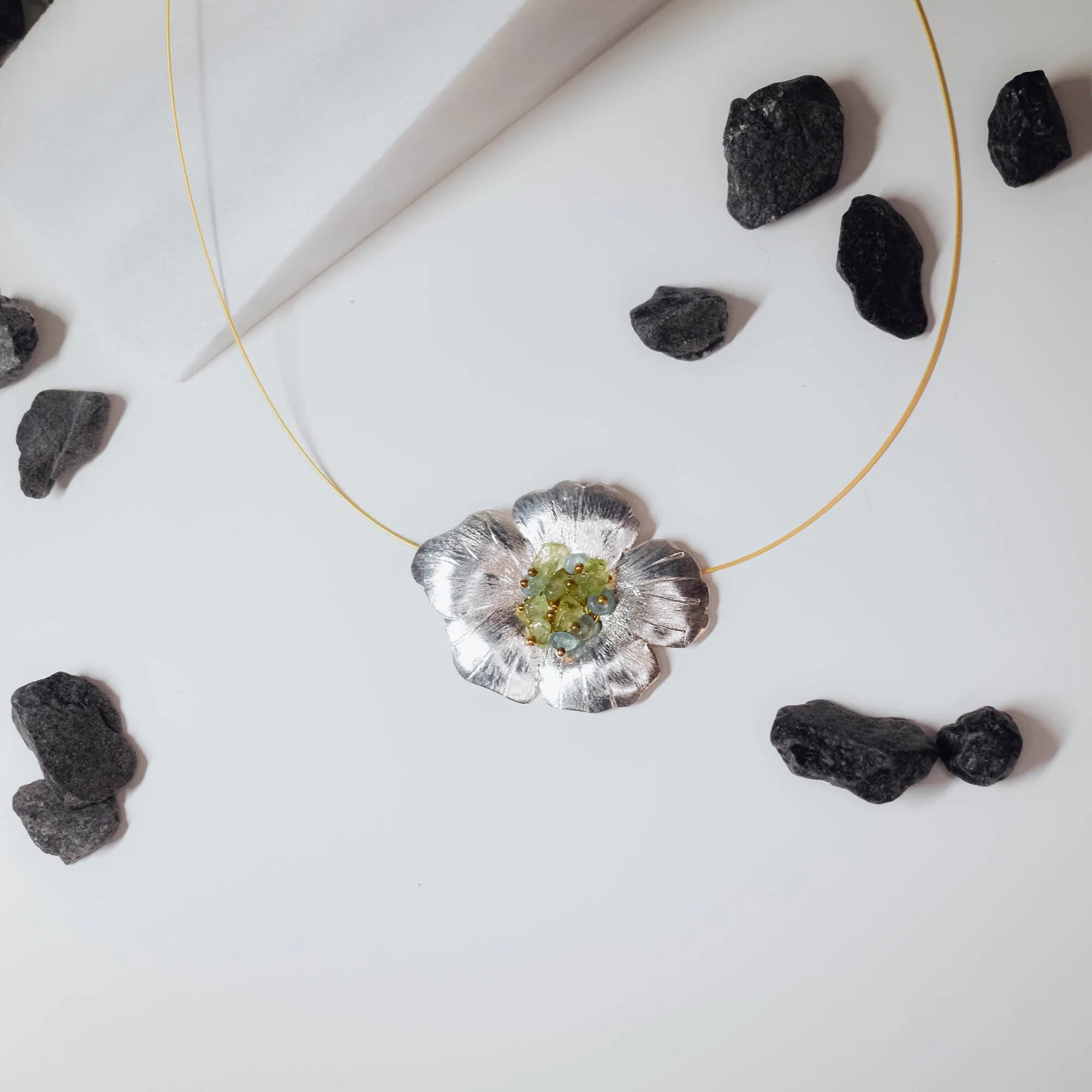 Large sculpted silver flower pendant with brushed petals and a raw green tourmaline and gold bead centre cluster, displayed on a gold wire choker on a white surface with black stones. Main image