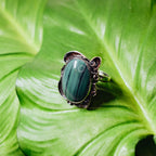 Side view of a silver malachite ring showing the ornate oxidised setting, the large oval malachite cabochon and the adjustable silver band on a green leaf background.