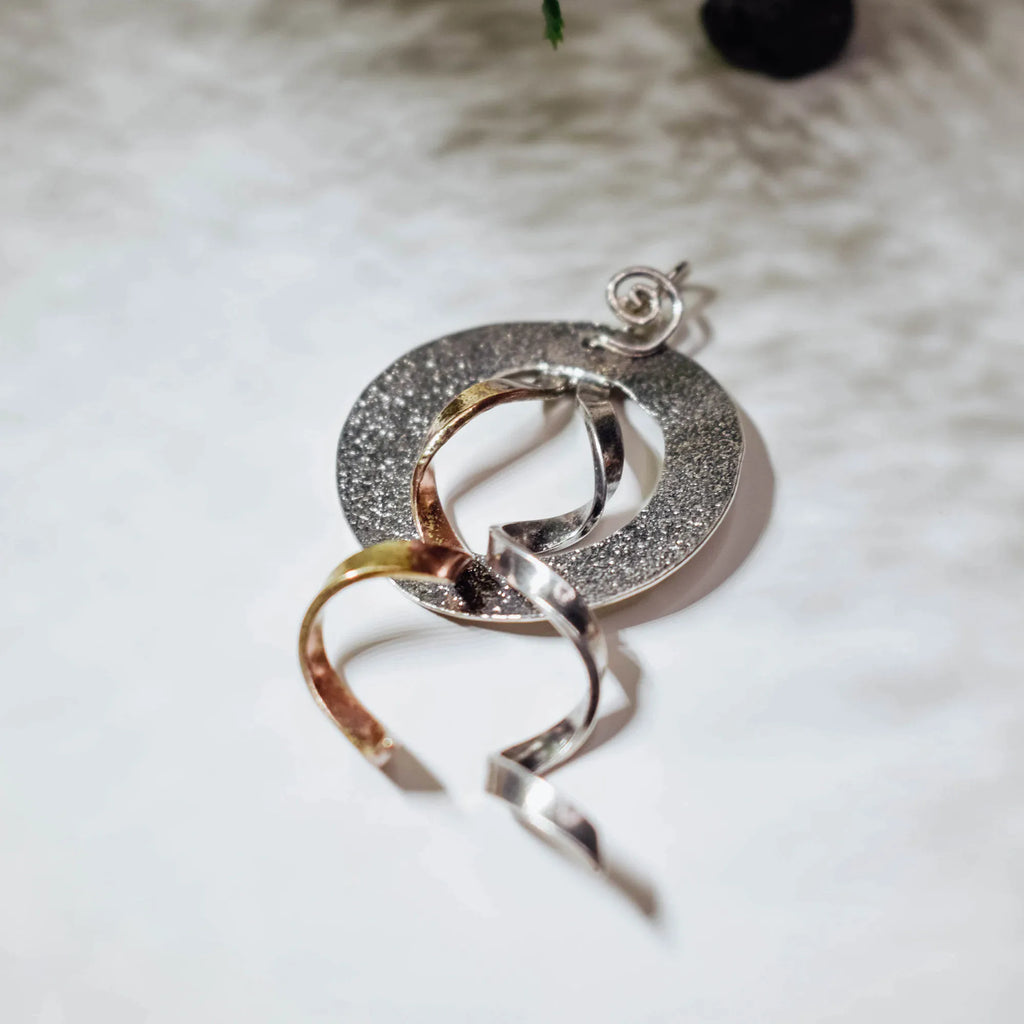 Side angle view of a textured circular silver pendant showing the two twisted ribbon elements in gold and polished silver crossing through the disc and flowing below.