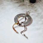 Side angle view of a textured circular silver pendant showing the two twisted ribbon elements in gold and polished silver crossing through the disc and flowing below.