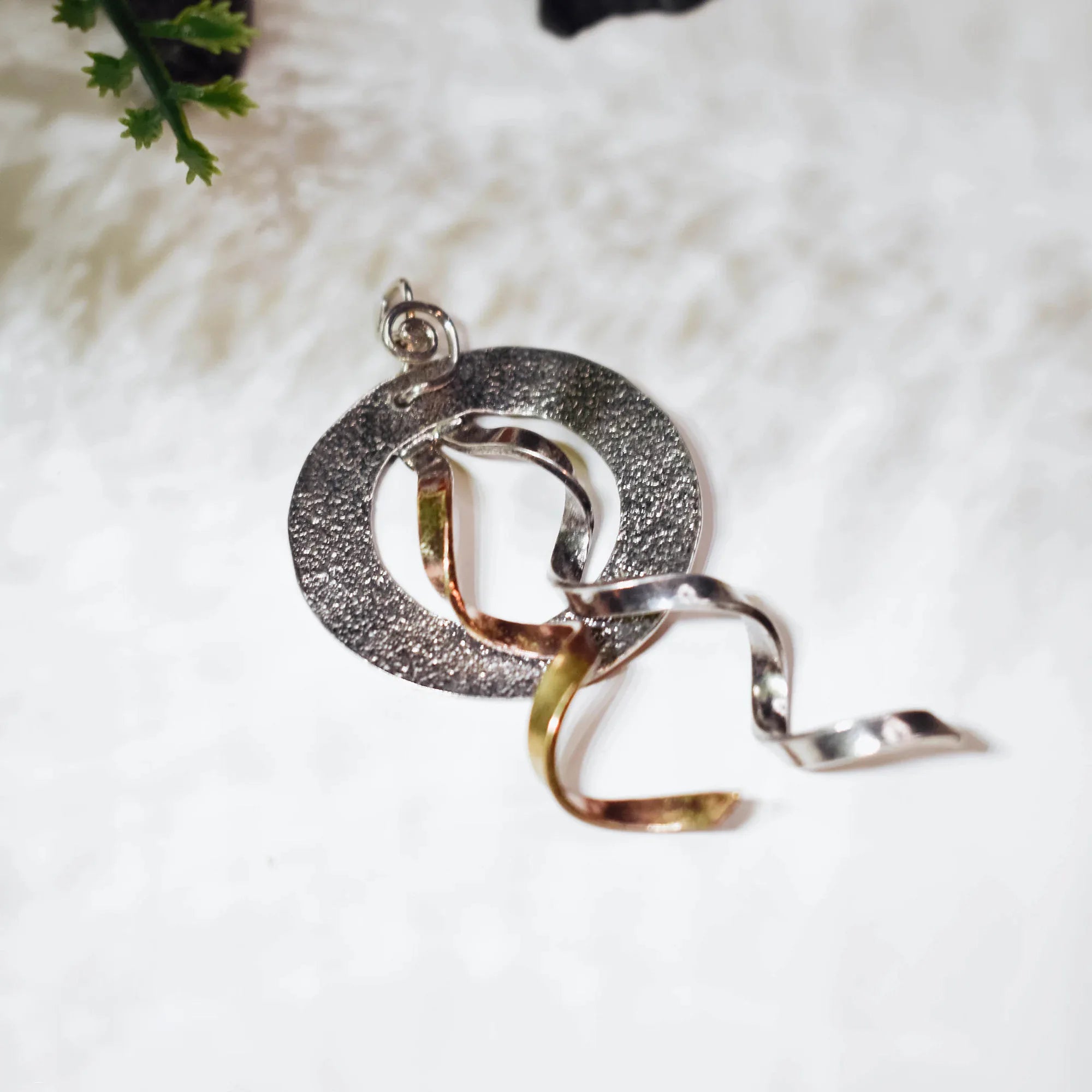 Top angle view of a textured circular silver pendant with spiral bail showing the two intertwining gold and silver twisted ribbon elements on a white surface.
