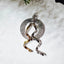 Sand-textured circular silver pendant with spiral bail and two intertwining twisted ribbon elements in gold and silver tones, displayed on a white fluffy surface.
