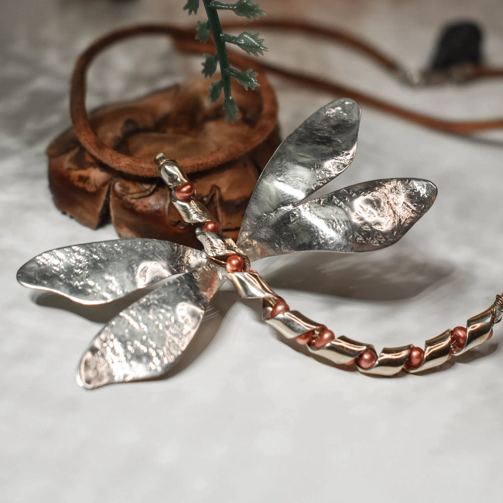 Close-up of a large silver dragonfly pendant showing the raw hammered wing texture and segmented body with copper-toned freshwater pearl accents on a leather cord.