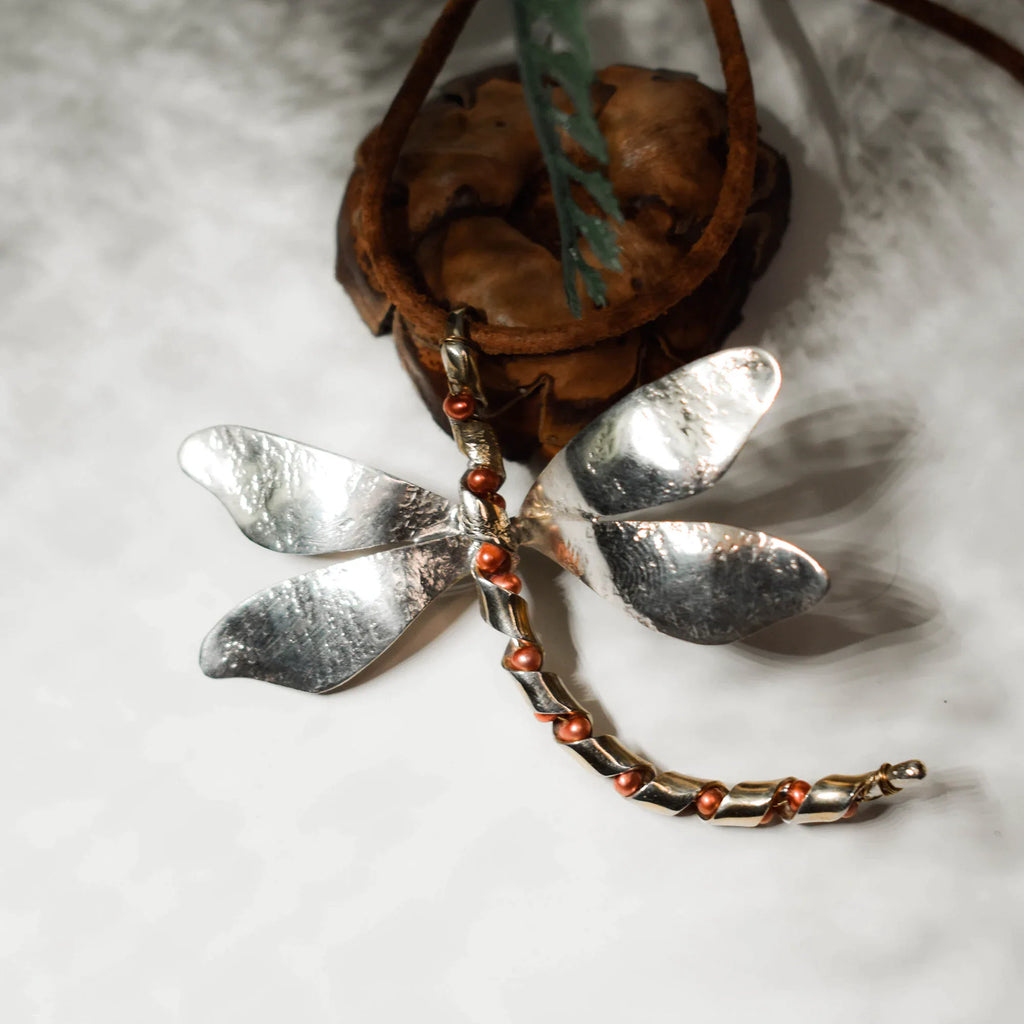 Large silver dragonfly pendant with four broad hammered wings and a segmented silver body with copper-toned freshwater pearl accents, displayed on a white surface with a leather cord.