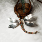 Large silver dragonfly pendant with four broad hammered wings and a segmented silver body with copper-toned freshwater pearl accents, displayed on a white surface with a leather cord.
