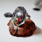 Side view of a silver hat-shaped ring showing the wide brim, domed crown and colourful gemstone cluster, displayed on a pine cone with dark stones in the background.