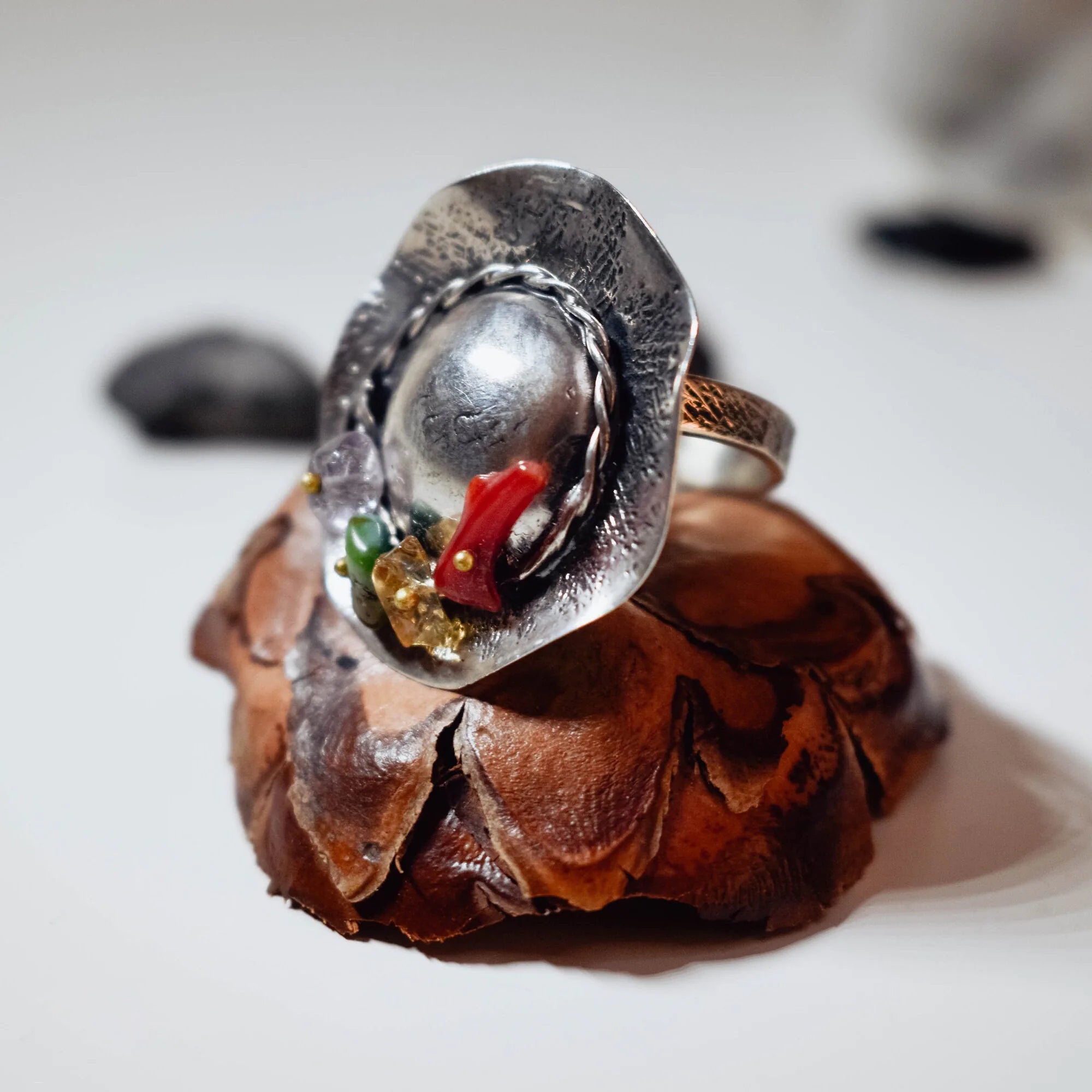 Side view of a silver hat-shaped ring showing the wide brim, domed crown and colourful gemstone cluster, displayed on a pine cone with dark stones in the background.