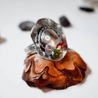 Silver hat-shaped ring with a domed crown, twisted wire hatband and a small cluster of red, purple and green stones, displayed upright on a pine cone with dark pebbles in the background.