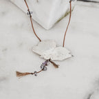 perspective view of a silver butterfly pendant with brushed engraved wings and purple amethyst chip drops on a brown tassel cord against a white marble surface.