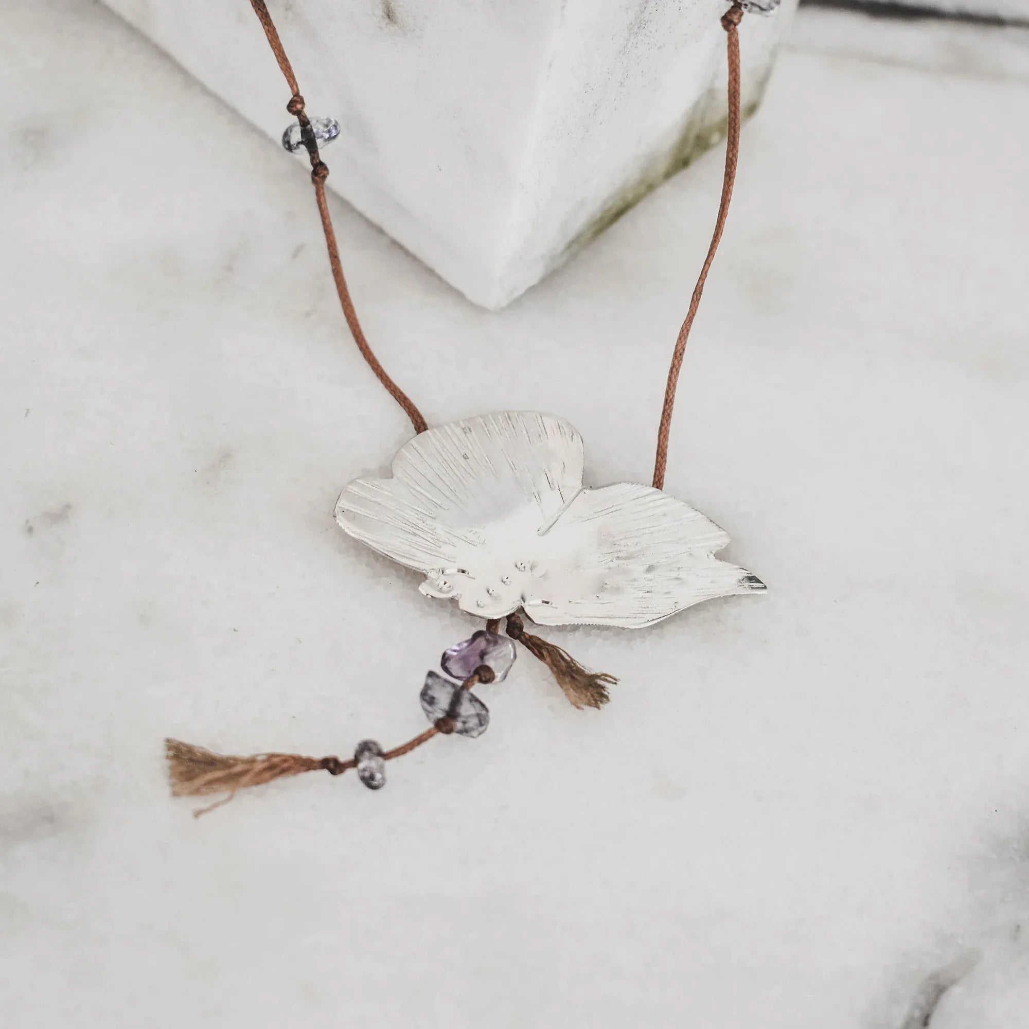 perspective view of a silver butterfly pendant with brushed engraved wings and purple amethyst chip drops on a brown tassel cord against a white marble surface.
