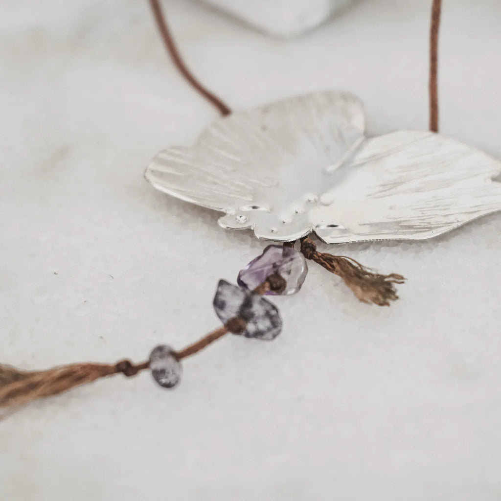 Close-up of a silver butterfly pendant showing the raw amethyst and purple gemstone chip drops hanging from the body with tassel cord ends and small knotted gemstone accents.