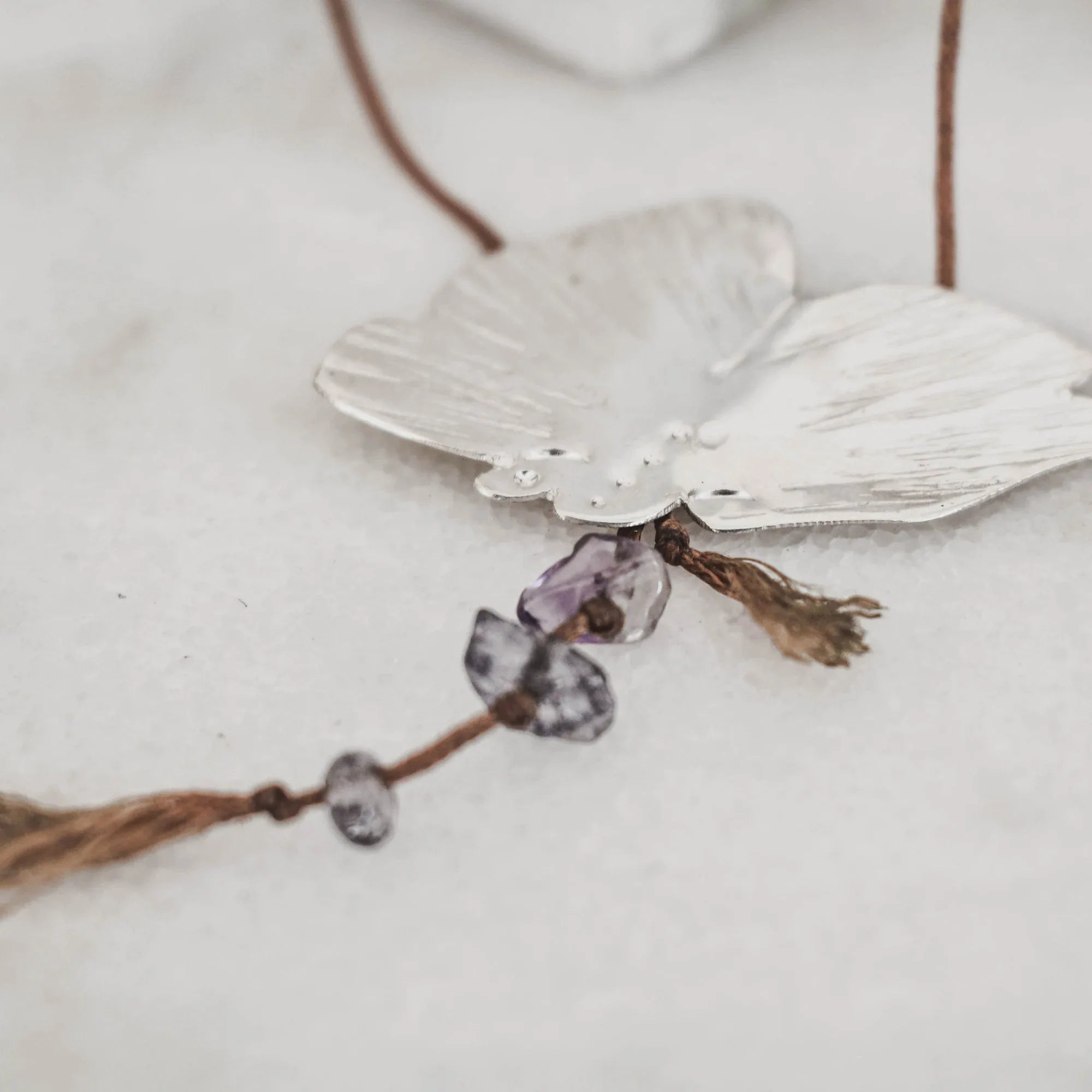 Close-up of a silver butterfly pendant showing the raw amethyst and purple gemstone chip drops hanging from the body with tassel cord ends and small knotted gemstone accents.