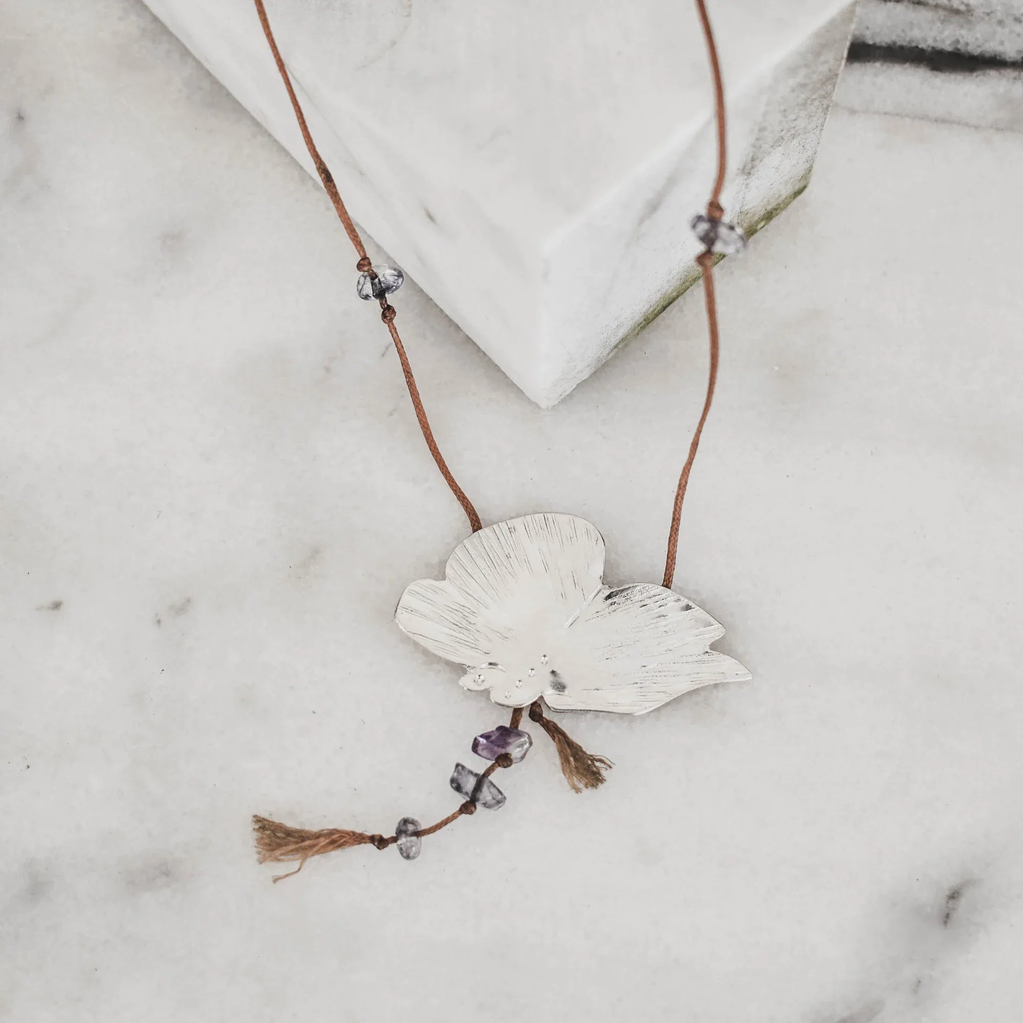 Silver butterfly pendant with engraved wings and raw amethyst drops hanging from the body, on a brown tassel cord with gemstone accents displayed on a marble surface.