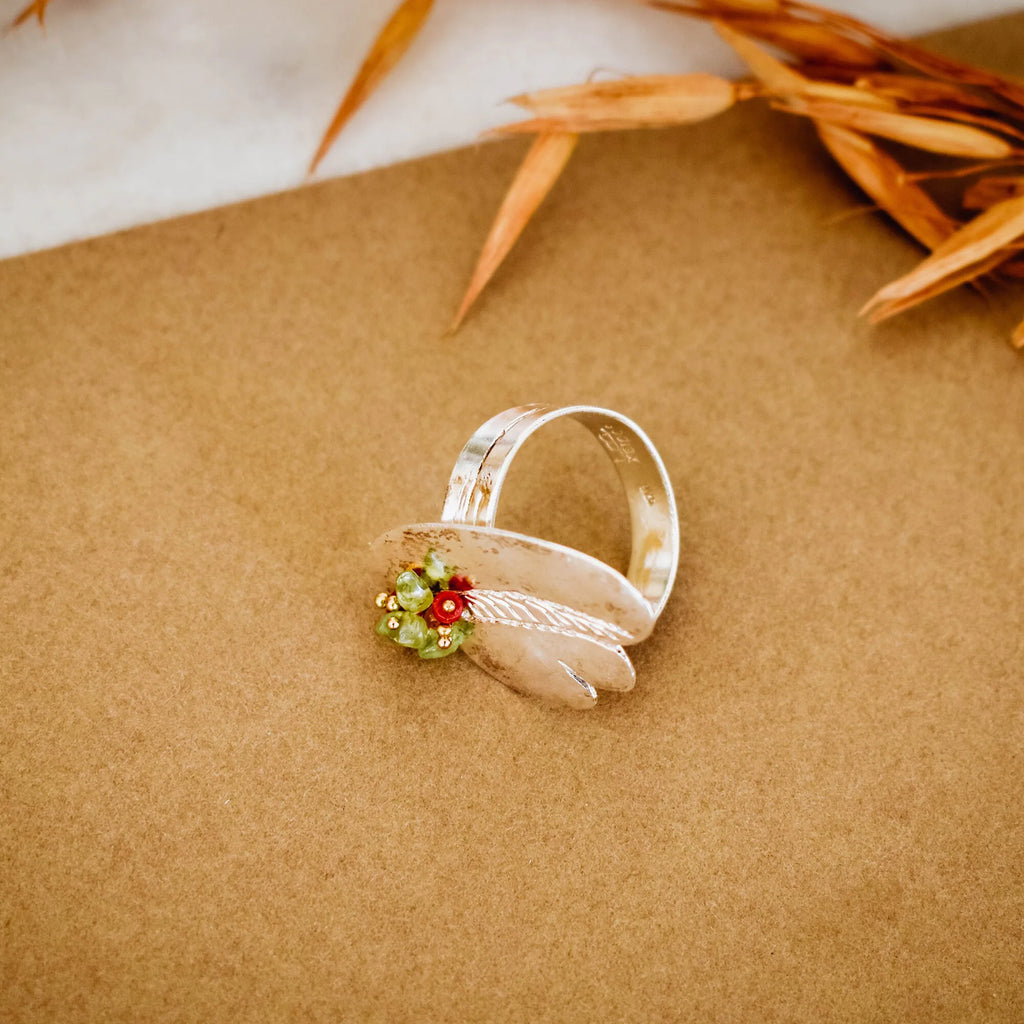 Silver ring with broad overlapping panels and a green and red gemstone cluster, lying flat on a kraft paper surface with dried botanical leaves in the background.