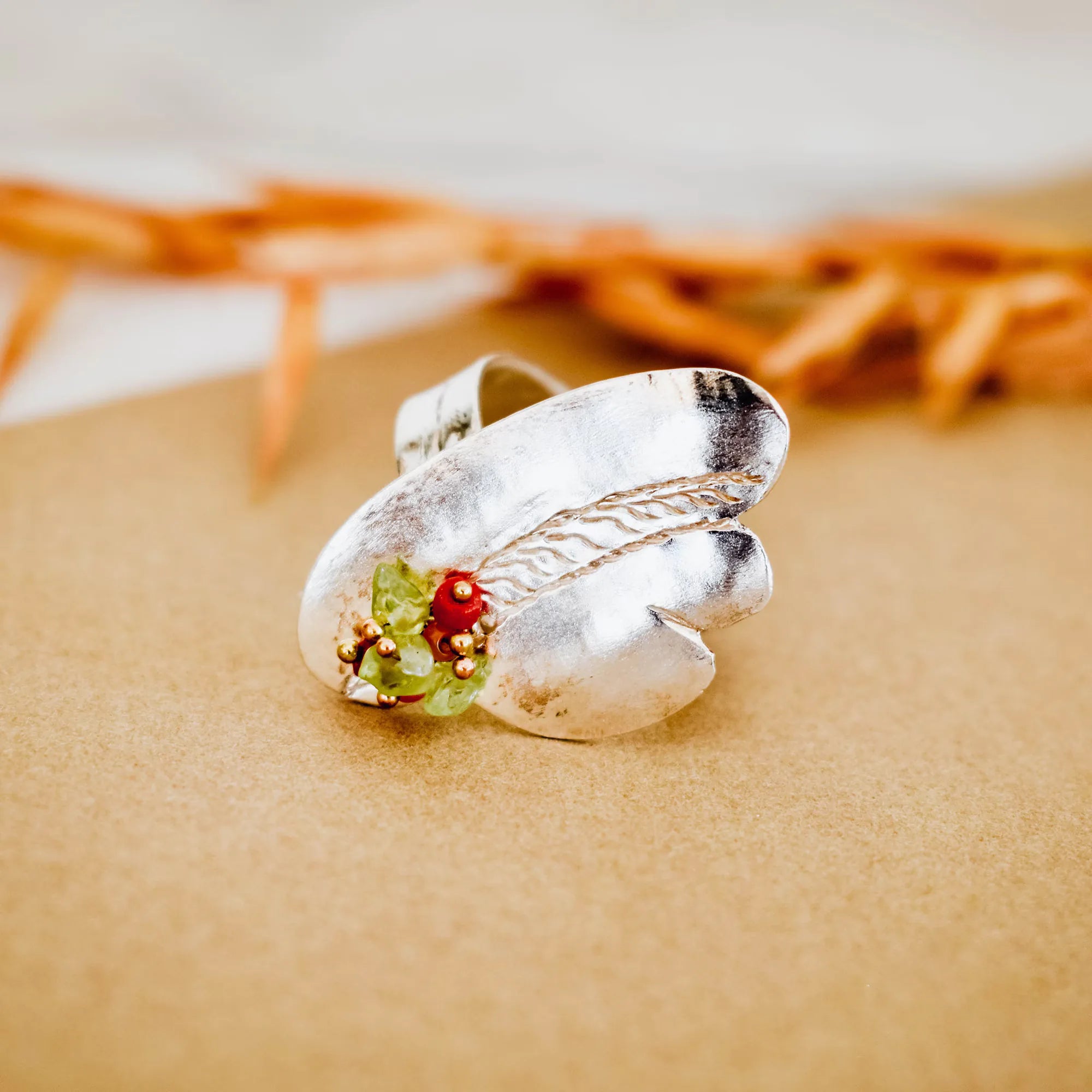 Wide silver ring with two overlapping curved panels and a small cluster of green and red stones with gold accent beads, resting on a kraft paper surface. Main image