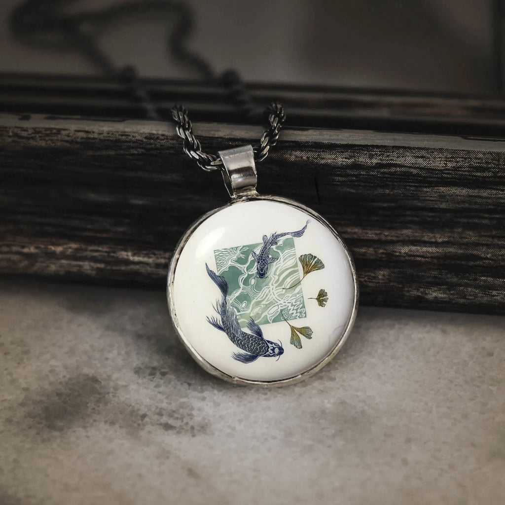silver pendant featuring blue koi fish swimming in stylized water with golden ginkgo leaves on a wooden background