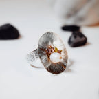 Side view of a large concave flower-shaped ring with radiating texture and a gemstone cluster at its heart, displayed on a white surface with dark pebbles in the background.