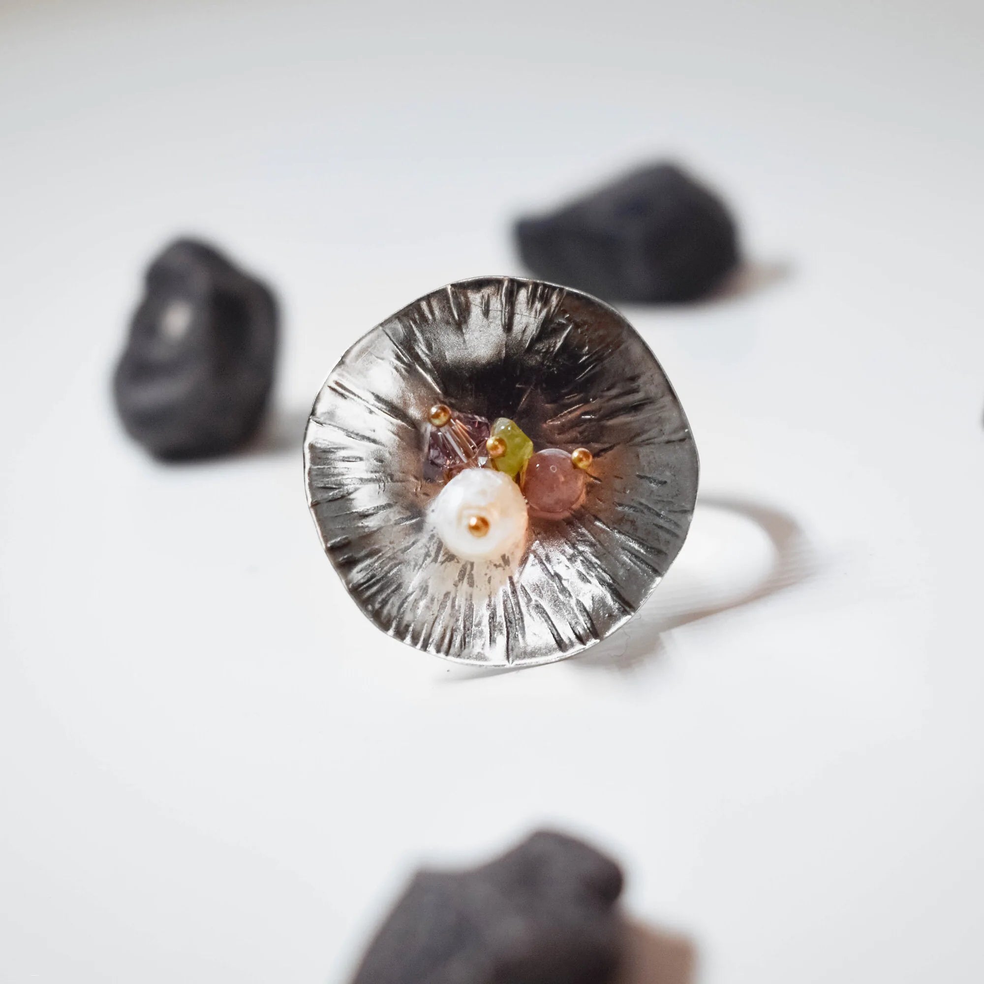 Front-facing view of a deeply oxidised concave flower ring with fine radiating lines and a delicate cluster of white, purple, green and pink stones nestled at the centre, with dark rocks surrounding it on a white surface.