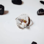Top-down view of a large concave flower-shaped ring with radiating petal lines and a small cluster of white, purple, green and pink stones with gold beads at the centre, surrounded by dark pebbles on a white surface.