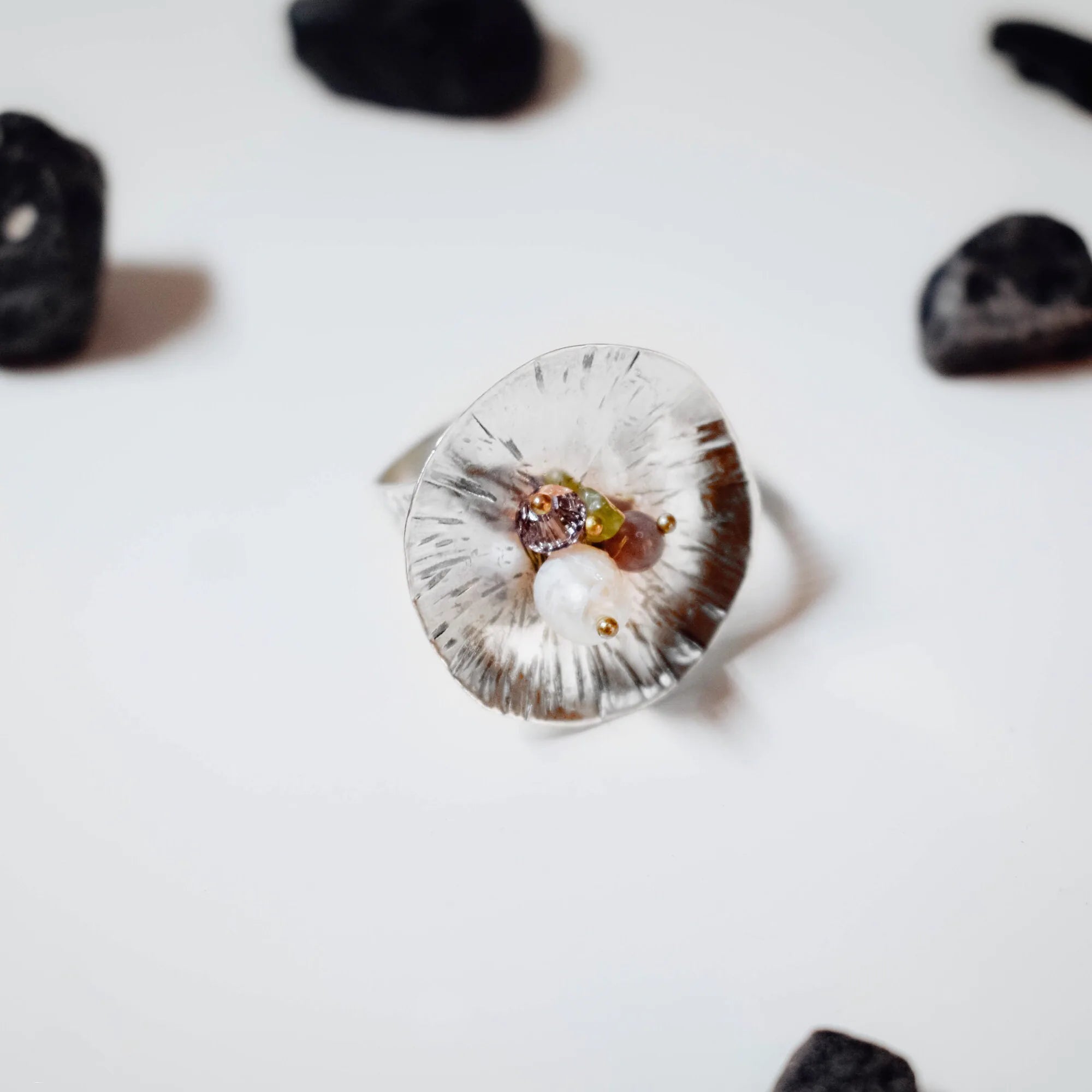 Top-down view of a large concave flower-shaped ring with radiating petal lines and a small cluster of white, purple, green and pink stones with gold beads at the centre, surrounded by dark pebbles on a white surface. Main image