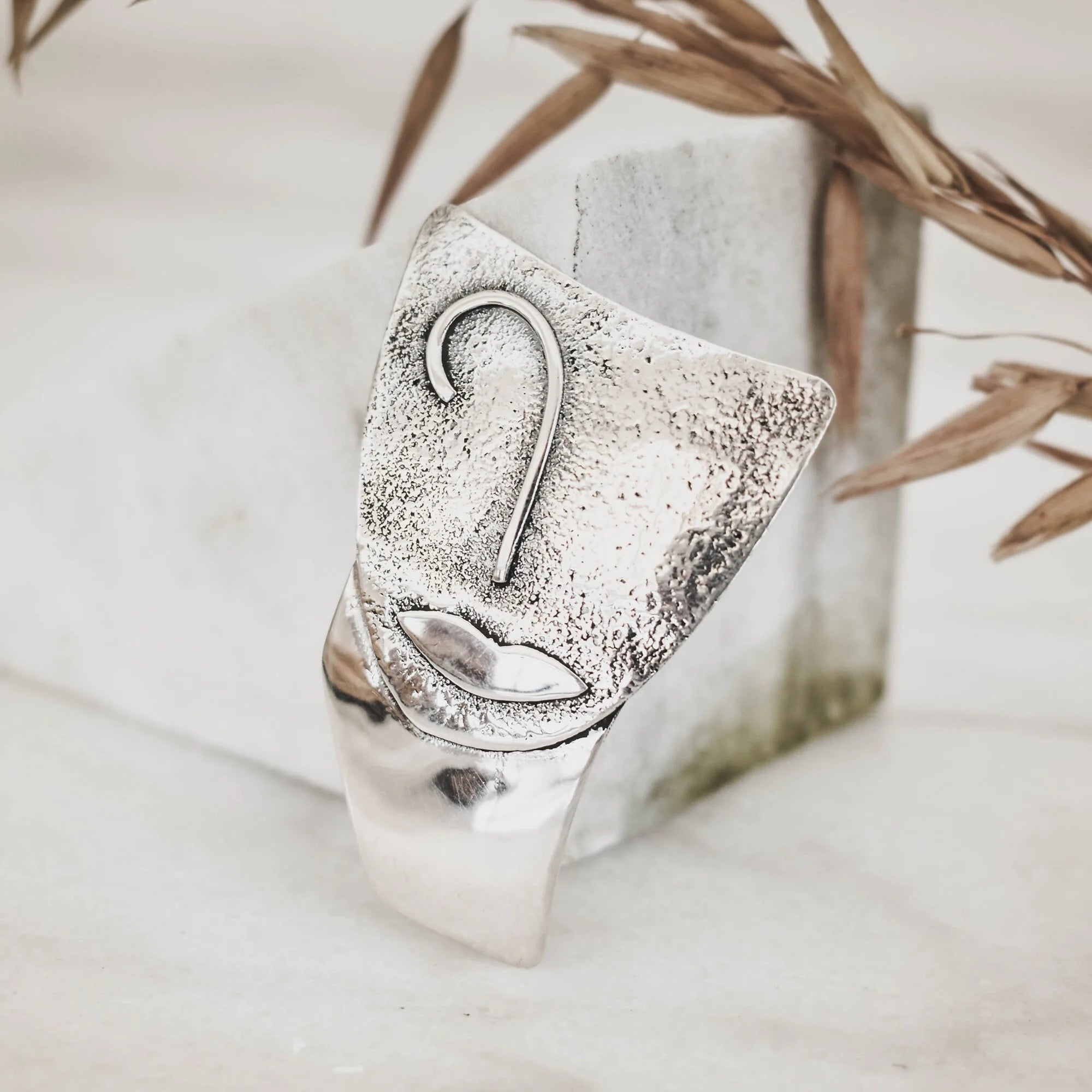 Abstract face silver pendant with heavily sand-textured upper surface showing a raised nose and sculpted lips, transitioning to a smooth polished lower section, displayed against a marble surface with dried grasses. Main image
