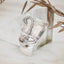 Abstract face silver pendant with heavily sand-textured upper surface showing a raised nose and sculpted lips, transitioning to a smooth polished lower section, displayed against a marble surface with dried grasses.